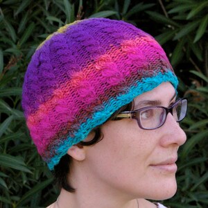 May include: A colourful knitted hat with a purple, pink, blue and brown colour scheme. The hat has a cable knit pattern and is being worn by a person with glasses.