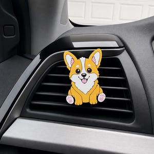 May include: A car air freshener clip featuring a cartoon illustration of a golden and white Corgi dog with large black eyes, pointed ears, and pink paw pads, attached to a black horizontal vehicle dashboard air vent slat.