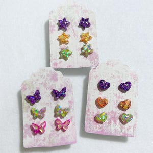 May include: Stud earrings in star, butterfly, and heart shapes. The earrings are glittery and come in purple, orange, green, and pink. Each pair is displayed on a floral patterned card.