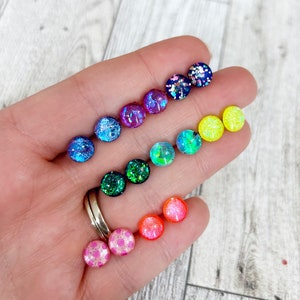 May include: A collection of colorful stud earrings in a variety of colors, including pink, orange, green, blue, and purple. Each earring is round and features glitter or a shimmering effect. The earrings are displayed in a hand.