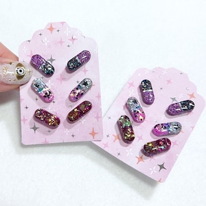 May include: Glittery capsule-shaped earrings in various colors, including purple, pink, and gold, are displayed on pink cards with star accents. The earrings are a unique accessory, perfect for adding a touch of sparkle to any outfit.