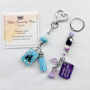 May include: Two keychains featuring rectangular resin charms. One is light blue with glitter and a black symbol, paired with a light blue tassel. The other is purple with glitter and white markings, accented by small purple flower and leaf charms.
