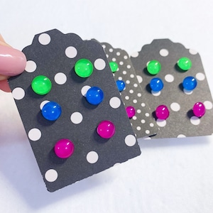May include: A collection of colorful stud earrings displayed on black tags with white polka dots. The earrings are in shades of green, blue, and pink. The earrings are small, round, and have a glossy finish.