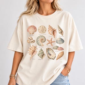Puede incluir: Una camiseta de manga corta color hueso con un estampado gráfico central de varias conchas y vida marina, incluyendo una vieira, un dólar de arena, un caracol, un nautilo, una estrella de mar, una almeja y una ostra, etiquetadas en cursiva.