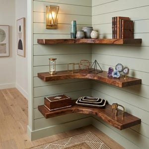 May include: Three tiered corner shelves made of natural dark wood with live edges, mounted on a light green shiplap wall. The shelves hold various decorative items including books, vases, an hourglass, geometric wire frames, crystals, and a box.