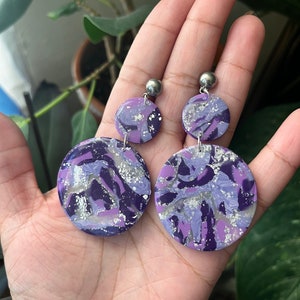 May include: A pair of dangle earrings with two round purple and silver marbled discs on each earring. The earrings have silver metal posts.