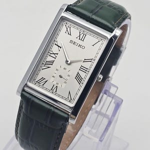 May include: A rectangular Seiko wristwatch with a silver-toned dial, black Roman numeral hour markers, and a small seconds sub-dial. The watch features a polished silver-tone case and a dark green leather strap with a textured crocodile-like pattern.