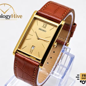 Può includere: Un orologio da polso rettangolare Seiko con quadrante dorato, lancette dorate e datario a ore sei. L'orologio presenta un cinturino in pelle marrone con motivo a coccodrillo e una cassa in metallo color oro lucido.
