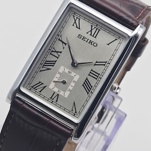 May include: A rectangular Seiko wristwatch with a silver-toned dial, black Roman numeral hour markers, and a small sub-dial for seconds. The watch features a polished silver-tone metal case and a dark brown leather strap with a textured finish.