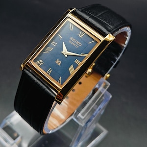 May include: A rectangular Seiko Quartz wristwatch featuring a gold-tone case, a deep blue dial with a textured wave pattern, gold Roman numeral hour markers, gold hands, and a black leather strap with a textured finish and a gold-tone buckle.