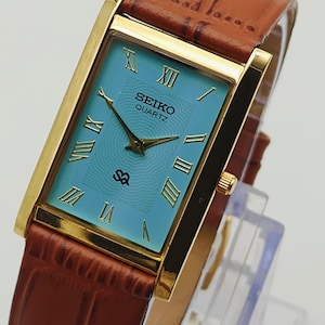 May include: A rectangular Seiko Quartz wristwatch featuring a gold-tone case, a light blue dial with a wavy texture, and gold Roman numeral hour markers. The watch includes gold hands and a brown leather strap with a textured crocodile-style pattern.