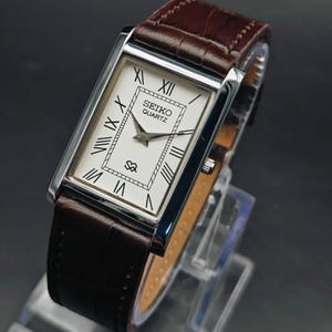 May include: A rectangular Seiko Quartz wristwatch featuring a white dial with black Roman numeral hour markers, a silver-tone metal case, and a dark brown leather strap with a textured finish. The watch face displays the Seiko Quartz logo.