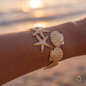 Puede incluir: Una pulsera de tono dorado que presenta una serie de dijes de metal conectados con formas de estrellas de mar y conchas estriadas. La pulsera tiene un acabado metálico pulido y está diseñada para rodear la muñeca con un patrón marino.