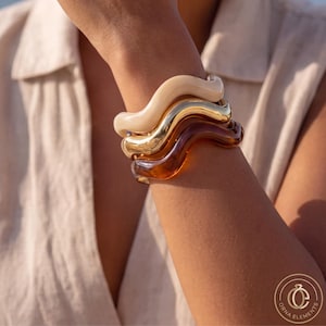 Puede incluir: Un conjunto de tres pulseras onduladas que incluye una banda de color crema suave, una banda metálica de tono dorado pulido y una banda de color ámbar marrón translúcido. Cada pulsera tiene una forma sinuosa y ondulada.