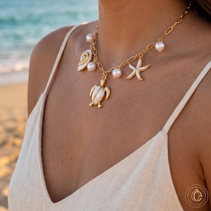Puede incluir: Un collar de cadena en tono dorado con un dije central de tortuga marina, un dije de estrella de mar y un dije de concha en espiral, todos con incrustaciones nacaradas blancas y perlas blancas redondas colgando de los eslabones.