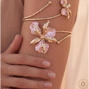 May include: A gold-toned metal arm cuff featuring a textured, open-ended band design. The piece is adorned with two floral motifs, each composed of metallic gold petals interspersed with soft, pale pink, iridescent enamel-like petal accents.