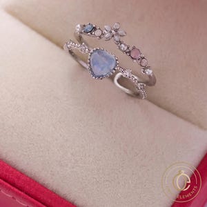 Puede incluir: Un conjunto de dos anillos plateados con una piedra azul claro en forma de corazón en la banda inferior y un motivo floral en la superior. La banda floral está decorada con pequeñas perlas blancas, piedras rosas y cristales transparentes, con un borde festoneado.