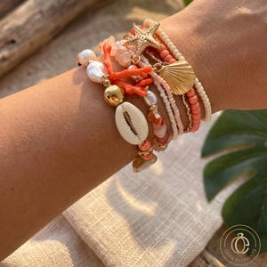Puede incluir: Un conjunto de pulseras de varias hebras con perlas blancas, cuentas de color coral y pequeñas cuentas de semillas color crema. El conjunto incluye dijes dorados con forma de estrella de mar y concha, junto con un acento central de cauri.
