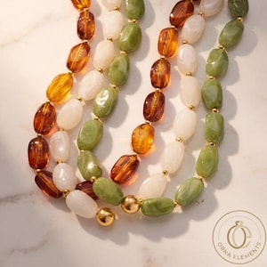 Puede incluir: Un collar de múltiples hilos con cuentas ovaladas en ámbar translúcido, blanco opaco y verde salvia jaspeado. Las cuentas están separadas por pequeños espaciadores de metal dorado, con cuentas esféricas doradas más grandes en los extremos.