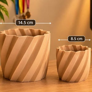 Op de afbeelding: Twee beige plantenbakken met een spiraalvormige, geometrische textuur. De grotere plantenbak heeft een diameter van 14,5 cm en een hoogte van 16 cm. De kleinere plantenbak heeft een diameter van 8,5 cm en een hoogte van 9,5 cm.