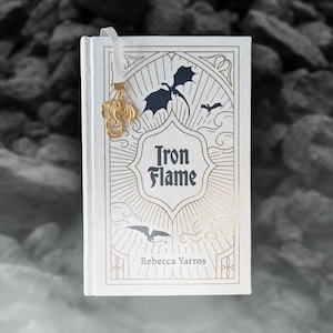 Peut inclure: Un livre relié blanc intitulé Iron Flame de Rebecca Yarros avec des détails en feuille d'or sur la couverture. La couverture présente des silhouettes de dragons noirs et un marque-page en ruban blanc attaché à un pendentif dragon en métal doré.