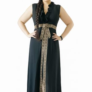 May include: A sleeveless black floor-length dress featuring a ruffled V-neckline and a wide, gold-toned patterned fabric belt at the waist. The belt includes a long, vertical decorative panel that extends down the center of the skirt.