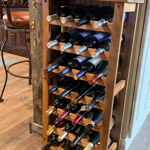May include: A tall, rectangular wooden wine rack with a natural brown finish, featuring six horizontal shelves designed to hold multiple glass wine bottles. The bottles are arranged in rows, showcasing various colored foil capsules and glass necks.