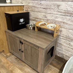 May include: A gray wooden cat litter box enclosure cabinet with two doors and a side entry hole. On top sits a black metal cat food storage bin with a white cat icon and the text "Cat Food," a silver bowl, and a wooden elevated double pet feeder.
