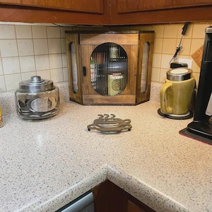 May include: A wooden corner cabinet with a central oval fluted glass door, a clear glass jar with a metal lid, a brown ceramic mug, and a dark metal trivet shaped like a coffee cup, all arranged on a speckled countertop.