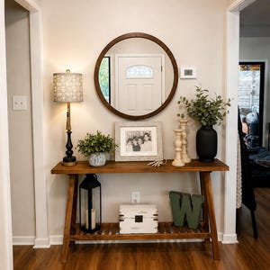 May include: A wooden console table with a lower shelf, featuring a round mirror, a lamp with a patterned shade, a framed photograph, two wooden candlesticks, a black vase with greenery, a black lantern, a white wooden box, and a green letter W.