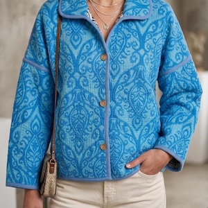 Könnte beinhalten: Eine gesteppte Jacke in leuchtenden Blau- und Hellblautönen mit einem sich wiederholenden Damastmuster. Die Jacke hat einen Kragen, lange Ärmel und eine Knopfleiste auf der Vorderseite mit drei braunen Knöpfen. Sie wird mit einer hellen Hose getragen.