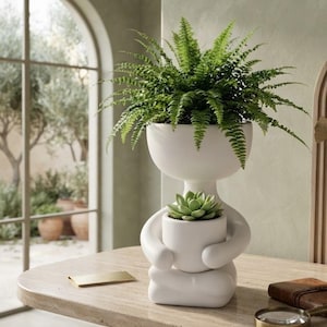May include: A matte white ceramic planter shaped like a stylized figure sitting in a cross-legged position. The figure holds a smaller cylindrical pot containing a green succulent, while a lush green fern grows from the head of the figure.
