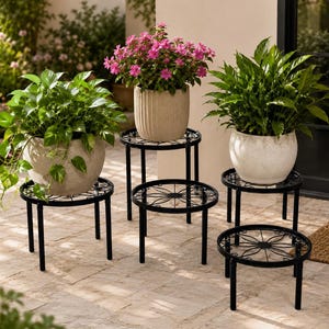 May include: A set of five black metal plant stands with circular tops featuring a decorative wire pattern and four straight legs. Three of the stands hold potted plants, including a trailing green vine and pink flowers in textured pots.
