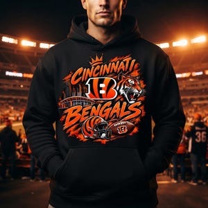 Puede incluir: Una sudadera negra con capucha que presenta un estampado gráfico del logotipo de los Cincinnati Bengals, una cabeza de tigre, un casco de fútbol americano, un balón y la silueta de un estadio en naranja y blanco con el texto Cincinnati Bengals.