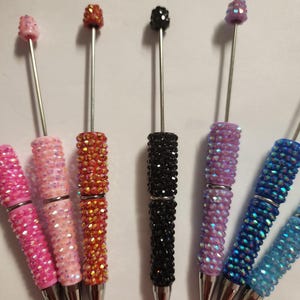 May include: A collection of six pens featuring barrels covered in small, reflective, multi-colored rhinestones. Each pen has a silver-toned metal tip and a thin, extended metal rod at the top, topped with a single matching rhinestone bead.