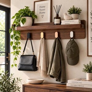 Può includere: Una mensola da parete in legno scuro con quattro ganci ovali neri, dorati, ramati e bianchi. La mensola ospita una pianta in vaso, una cornice con la scritta Home Sweet Home, un diffusore, una statuetta di uccello e due libri The Kinfolk Home.