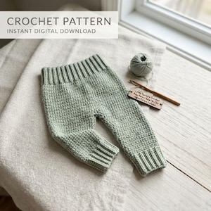 May include: A pair of sage green crocheted baby pants with a ribbed waistband and ribbed cuffs. Next to the pants is a ball of matching yarn, a wooden crochet hook, and a small wooden tag labeled Little Sprouts Crochet Baby Pants 0-12M.