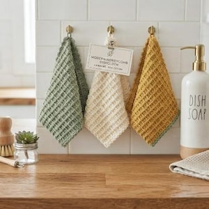Könnte beinhalten: Drei quadratische Spültücher mit strukturiertem Wabenmuster in Salbeigrün, Creme und Senfgelb, die an goldenen Haken hängen. Ein weißer Keramik-Seifenspender mit der Aufschrift "DISH SOAP" und ein gefaltetes cremefarbenes Spültuch sind ebenfalls zu sehen.