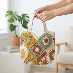 May include: A crochet tote bag constructed from multiple square motifs featuring floral patterns in shades of mustard yellow, sage green, terracotta, and cream. The bag includes two integrated handles and a V-shaped top opening.