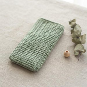 May include: A rectangular sage green crochet pouch featuring a consistent ribbed texture throughout the surface. The item is positioned next to a single small wooden bead and a dried eucalyptus stem with several rounded leaves.