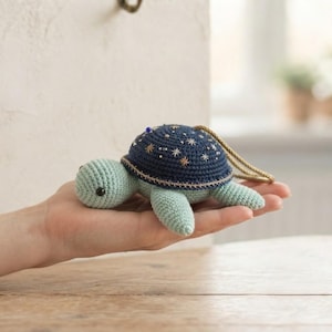 May include: A small, crocheted turtle toy with a light mint green body and a dark navy blue shell. The shell is decorated with embroidered silver star patterns and a single blue bead. A braided tan cord is attached to the top of the shell.