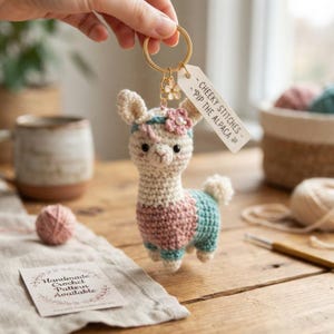 May include: A crochet alpaca keychain featuring a cream-colored head, a pink midsection, and a teal lower body. The alpaca has a teal headband with a small pink flower and a white tag reading CHEEKY STITCHES - PIP THE ALPACA attached to a gold ring.