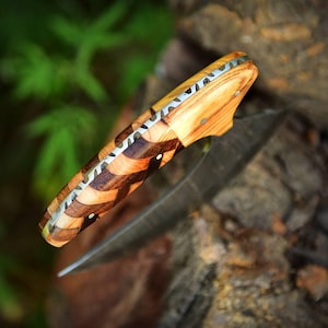 May include: A folding pocket knife featuring a handle crafted from alternating light and dark wood segments arranged in a chevron pattern, accented with a metallic spine and small circular rivets along the side of the wooden grip.