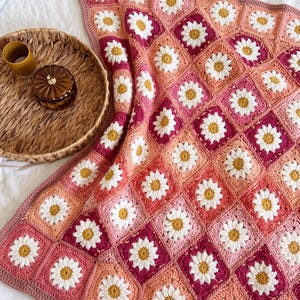Könnte beinhalten: Eine handgefertigte Häkeldecke aus quadratischen Motiven mit weißen Gänseblümchen und gelben Mitten. Die Quadrate sind in einem Farbverlauf aus Rosa-, Magenta- und Pfirsichtönen angeordnet und mit einem gewellten Rand versehen.