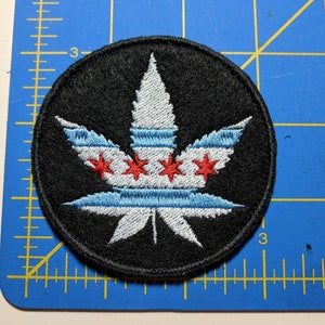 Könnte beinhalten: Ein runder schwarzer Patch mit einem weißen Marihuana-Blatt-Design. Das Blatt ist in Blau und Rot umrandet, was an die Flagge von Chicago erinnert.