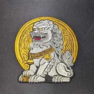 Puede incluir: León blanco bordado con detalles dorados en un parche circular. El león está sentado con la cabeza girada hacia un lado y la boca abierta. El parche está rodeado por un círculo dorado con un diseño decorativo.