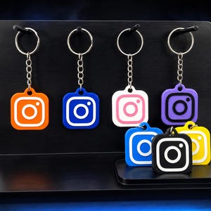 May include: A collection of square keychains featuring the Instagram logo in various colors including orange, blue, white, purple, yellow, and black. Each keychain is attached to a silver metal split ring by a short silver chain link connector.