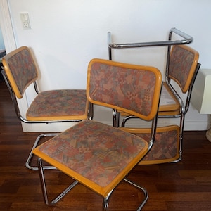Peut inclure: Un ensemble de trois chaises cantilever dotées de cadres en métal chromé poli, avec des dossiers et des assises en bois recouverts d'un tissu à motifs géométriques multicolores. Les chaises présentent une base tubulaire en métal.