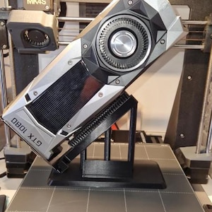 May include: A silver and black GTX 1080 graphics card mounted on a black plastic display stand. The card features a prominent circular fan, angular metallic casing, and visible cooling fins. The text GTX 1080 is printed on the side of the unit.