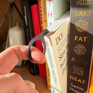 Puede incluir: Un sujetador de páginas de libro circular de plástico gris oscuro con una lengüeta plana y extendida que se inserta entre las páginas. El dispositivo cuenta con un mango en forma de anillo diseñado para sostenerse con un dedo.